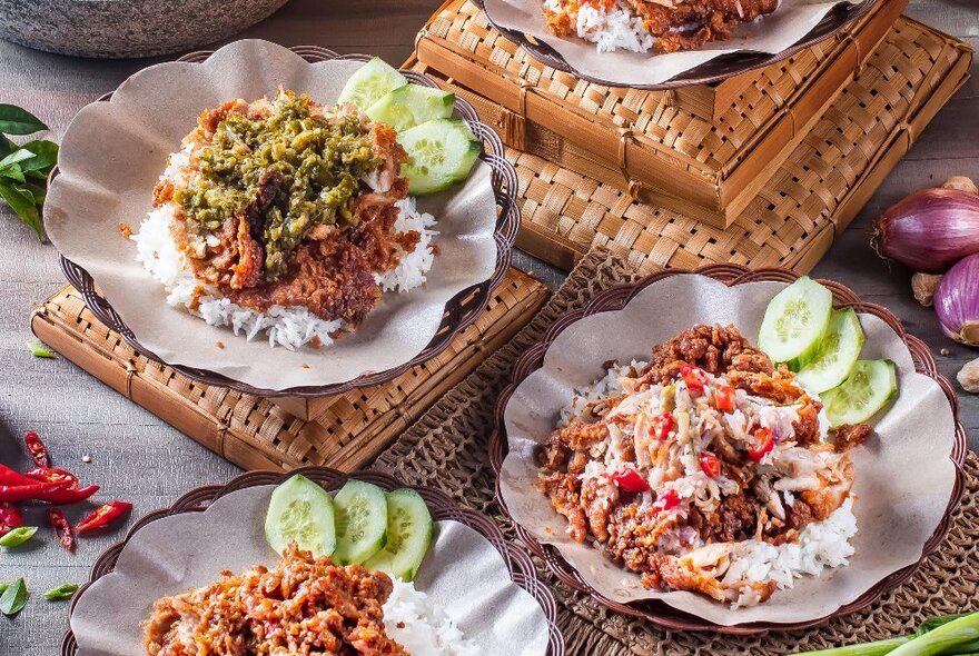 Bowls of delicious Ayam Geprek, an Indonesian staple consisting of smashed fried chicken served with rice and sambals, with a cucumber garnish.