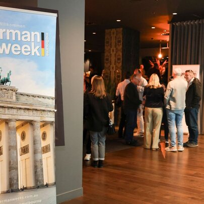 A room with people mingling and a large poster of the Brandenburg Gate that says German Week. 