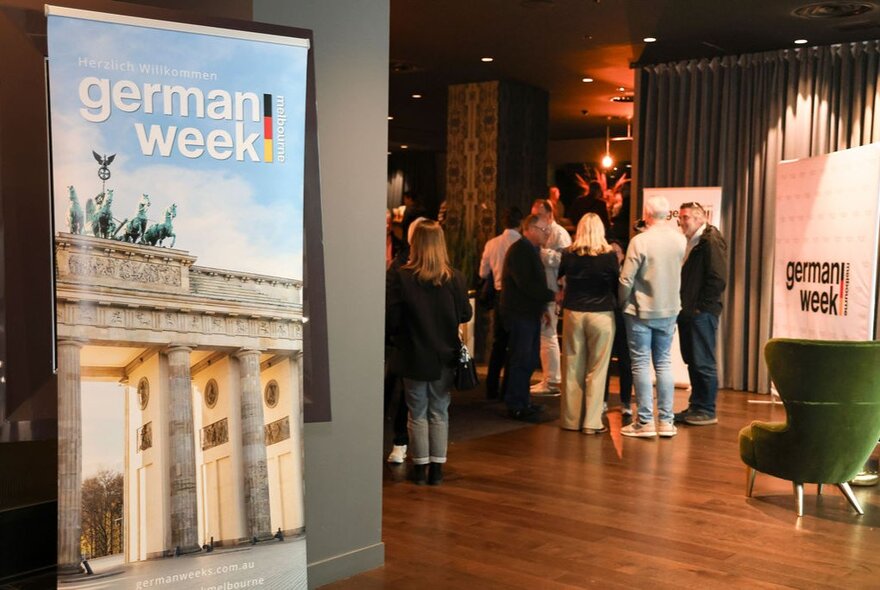 A room with people mingling and a large poster of the Brandenburg Gate that says German Week. 