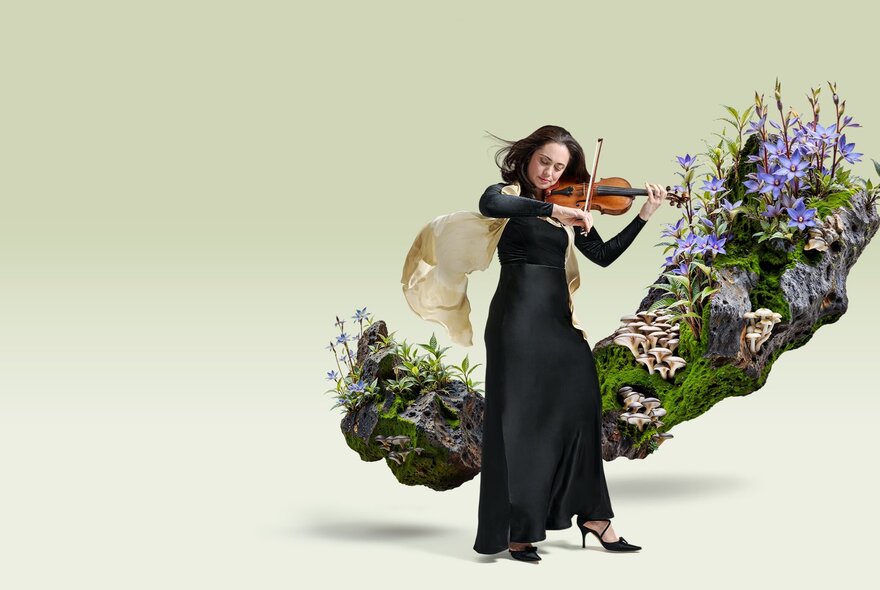A violinist wearing a long black dress, playing a violin, standing in front of a rocky outcrop of green mossy and blue flowers which appears to float against a pale background.