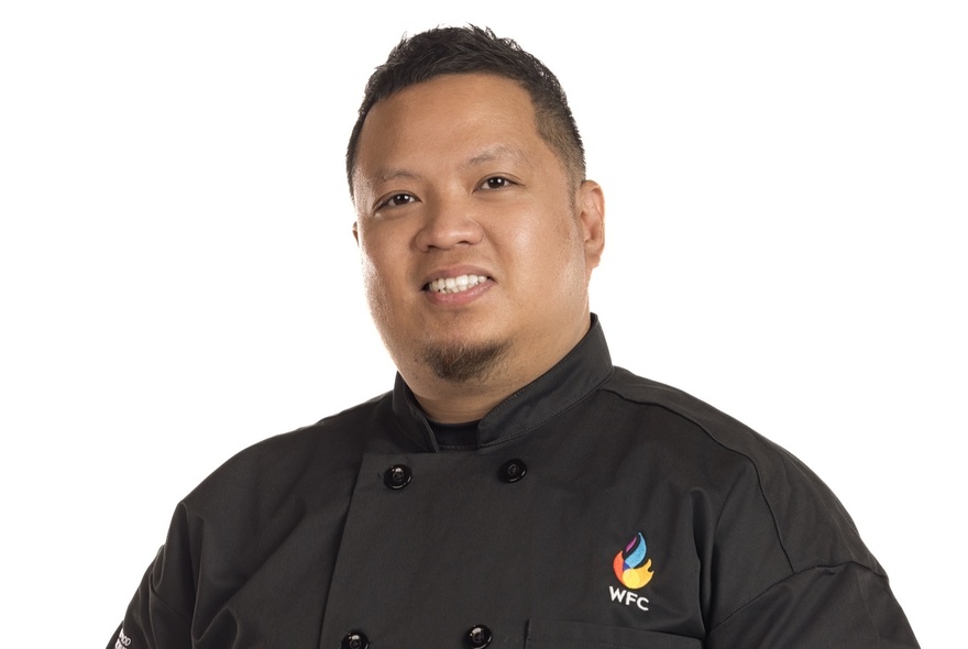 Portrait of a male chef with short black hair wearing a black chef's shirt.