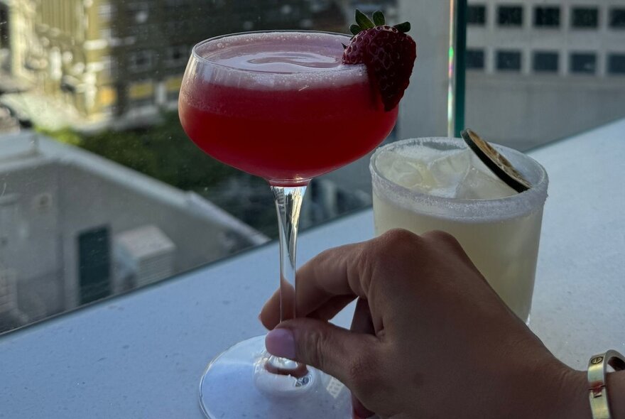 Hand holding a red cocktail at a rooftop bar with city views.