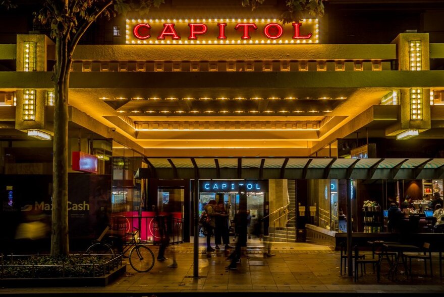 Outside an art deco theatre at night with glowing lights and a red illuminated sign saying Capitol