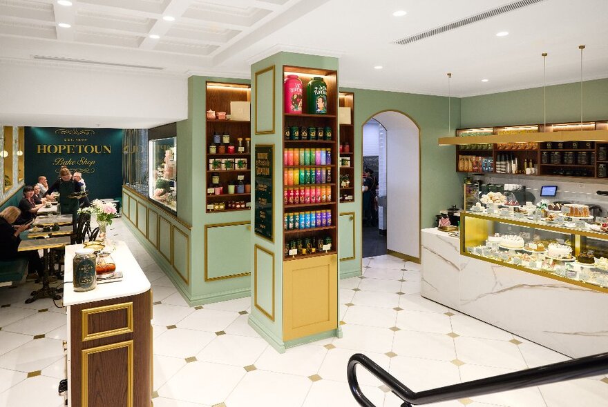 Cafe interior showing a service counter, products on display on shelves, a tiled floor, and people seated at small tables towards the back of the shop.