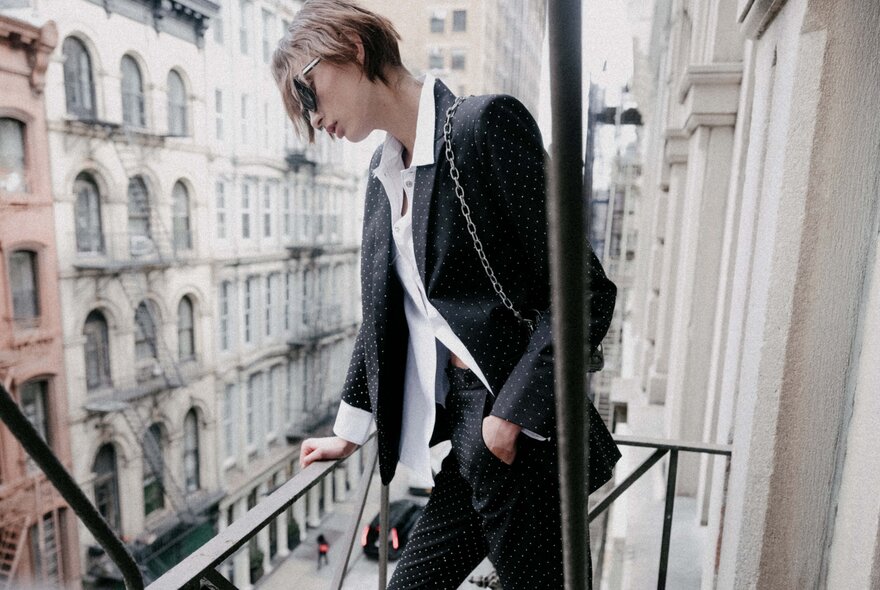 Model wearing a suit and sunglasses, standing looking down from an apartment building balcony.
