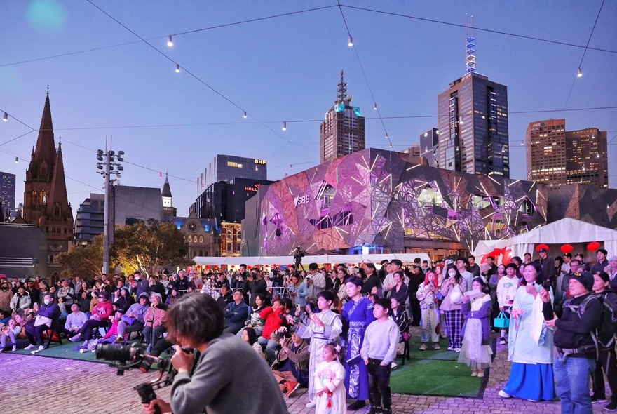 An image of Federation Square at dusk with people watching a performance (not visible).
