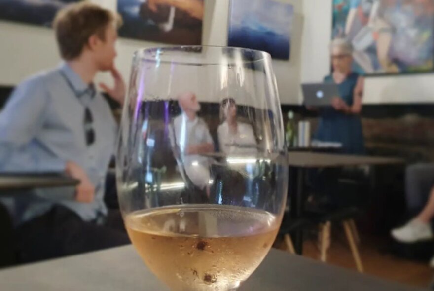 Looking through a glass of white wine to patrons in a restaurant, listening to someone reading. 