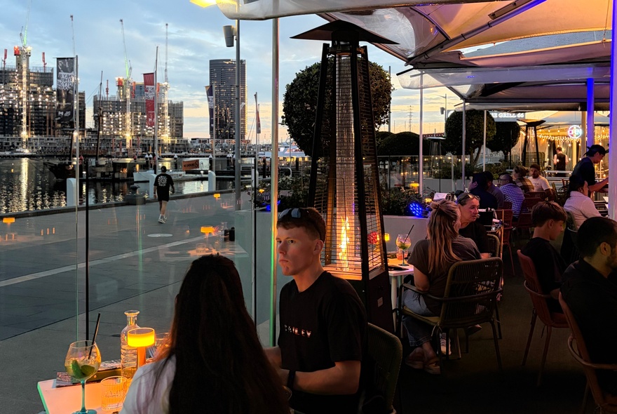 Diners seated outside under awnings overlooking Docklands harbour with city buildings in the background.