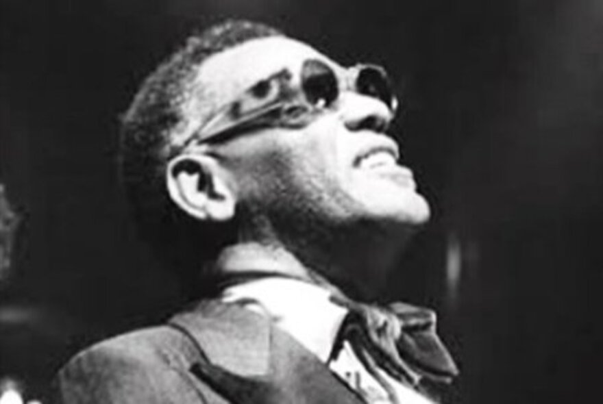 Old black and white photograph of Jazz musician, Ray Charles, wearing a suit and cravat and sunglasses, looking up.