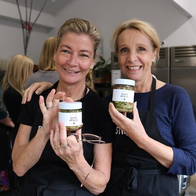 Two smiling woman holding up jars of sauerkraut in a commercial kitchen setting.