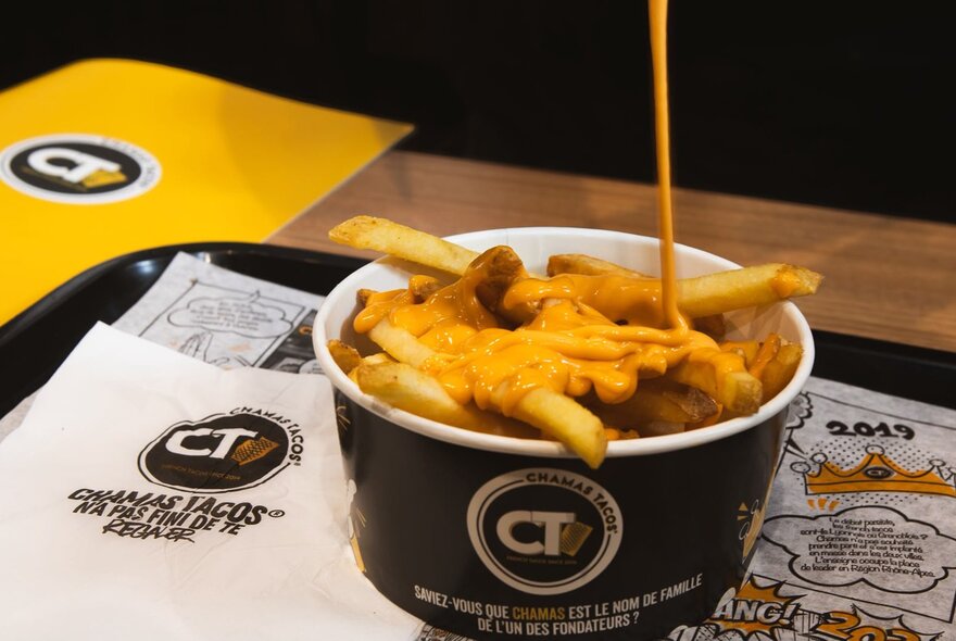A bowl of French fries topped with melted cheese on a tray inside a fast food restaurant.