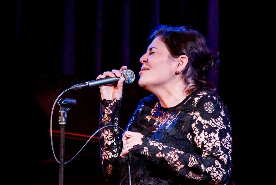 A woman in a sheer black lace top singing expressively into a microphone, 