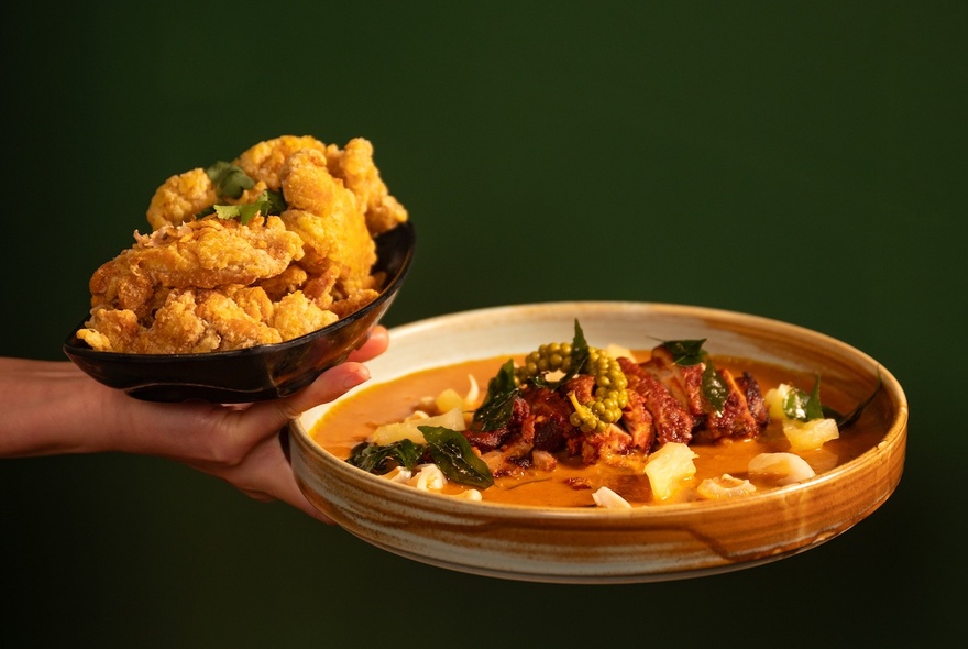 A person's hand holding a dish of Asian fried chicken and also a red duck curry in an earthenware bowl.