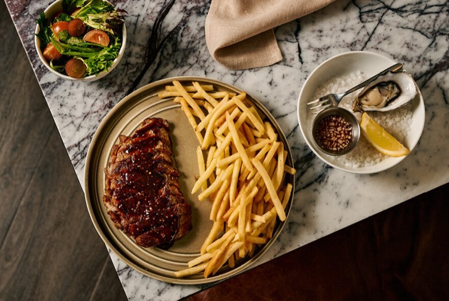 A selection of dishes on a marble table seen from above, including steak and fries, an oyster with lemon, and a garden salad.