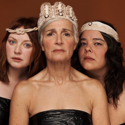 Three women dressed in strapless gowns with head bands or crowns looking mournful. 