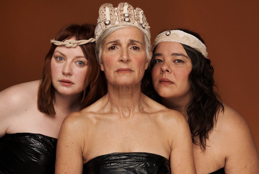 Three women dressed in strapless gowns with head bands or crowns looking mournful. 