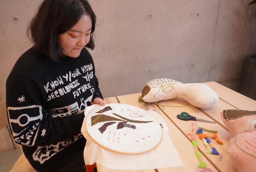 A person seated at a table doing embroidery, with soft fabrics, scissors and thread on the table.