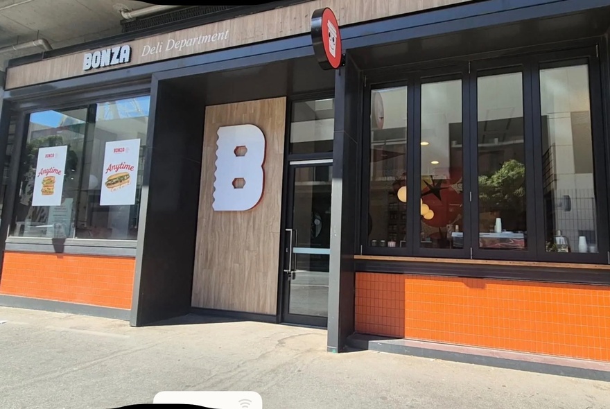The storefront of the deli and sandwich shop Bonza, featuring a large white 'B' logo on a wooden panel door and orange lower wall sections.