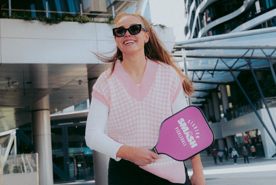 A person smiling, wearing sunglasses and holding a pink pickleball paddle outside the front of Marvel Stadium.