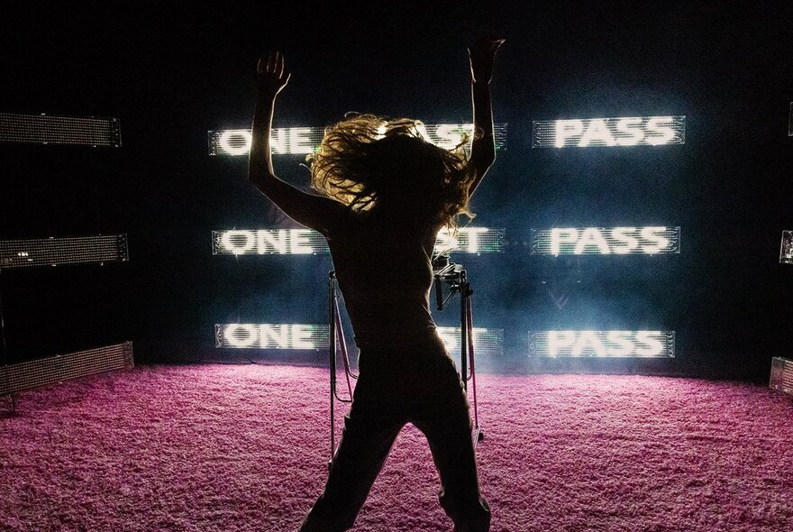 A silhouette of a person dancing on a stage with a pink floor, in front of screens displaying the words "ONE LAST PASS"