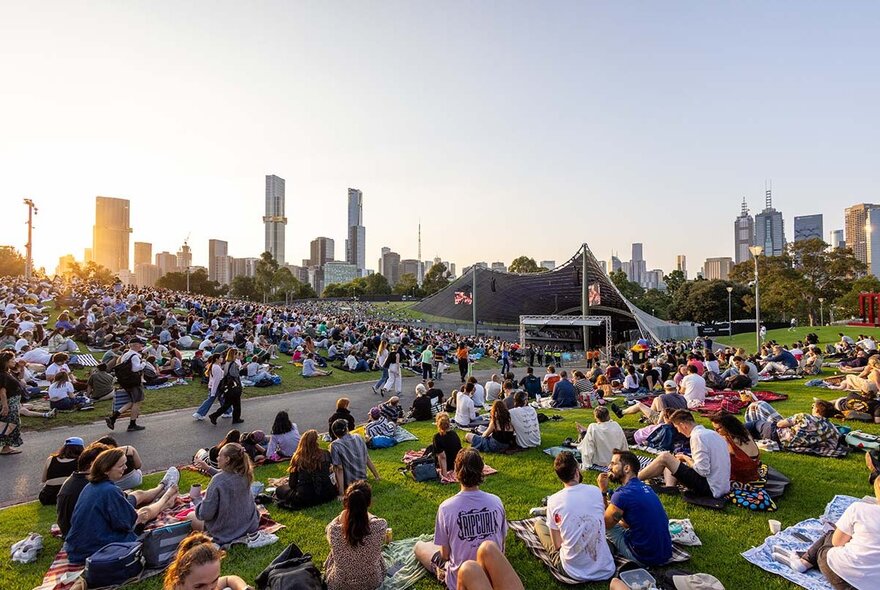 A large crowd of people spread out over the lawn at the Sidney Myer Music Bowl at dusk.