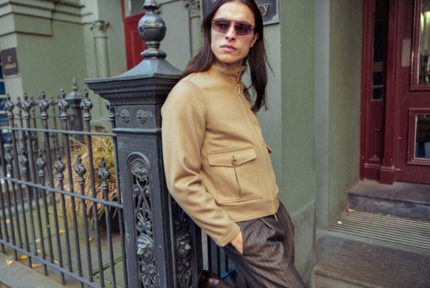 Side angle of man leaning up against Victorian style fence wearing camel coloured jacket.