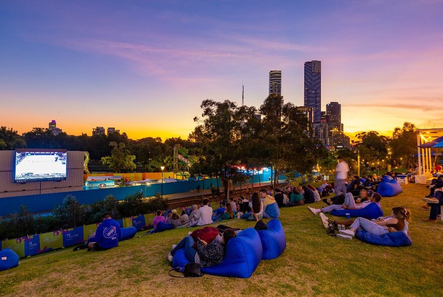 A crowd of people are watching tennis on a big screen on a hill at sunset. 