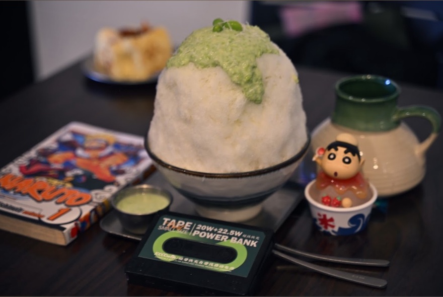 A very large bowl of kakigori, piled hight with matcha topping on a tablw with comics and a a small novelty creature. 