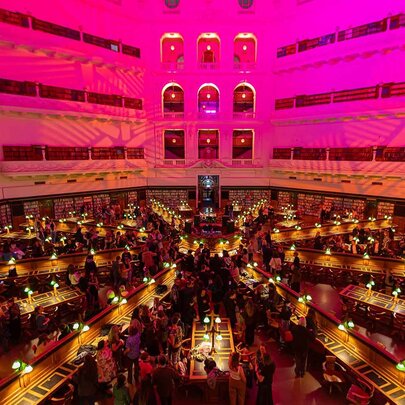 The Dome reading room of State Library Victoria showing the upper level balconies, and rows of desk fanning out from the centre, all bathed in a pink light.