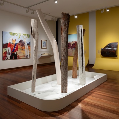A gallery space with paintings displayed on one white and one yellow wall, plus a series of totems or tree trunks displayed in a white stand in the middle.