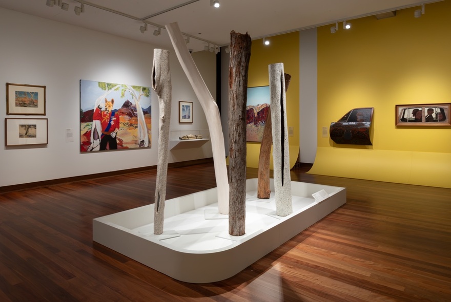 A gallery space with paintings displayed on one white and one yellow wall, plus a series of totems or tree trunks displayed in a white stand in the middle.