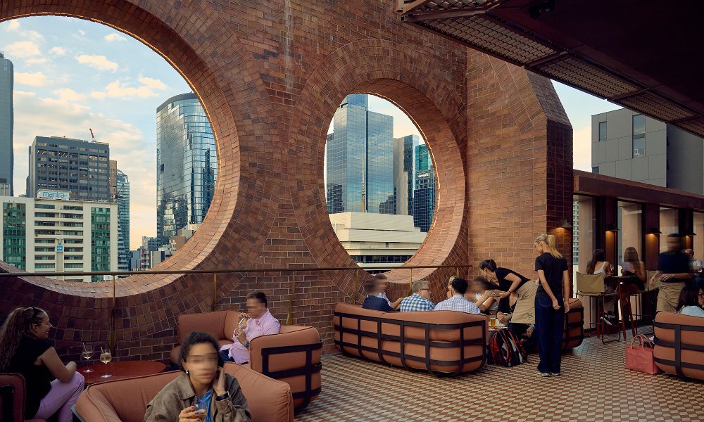 people sitting on couches on a stylish rooftop bar, with cutout circle windows in the brick walls, overlooking the city.