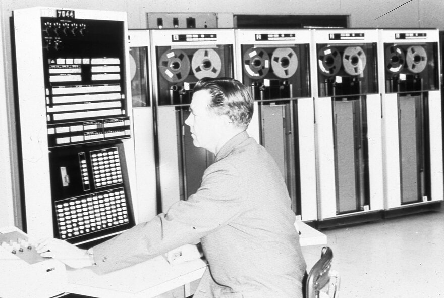 A black and white photo of a man sitting in front of one of the earliest computers.