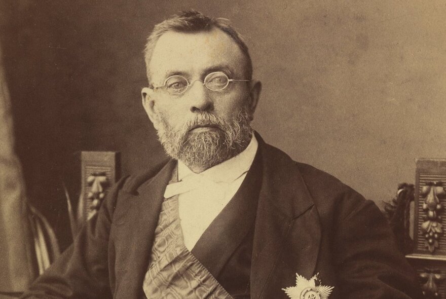 Sepia toned vintage image of Baron Ferdinand Von Mueller, with small round spectacles and wearing a sash over his white shirt and jacket.