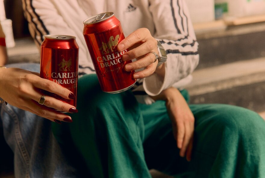 Two individuals' hands, each holding a red can of beer.