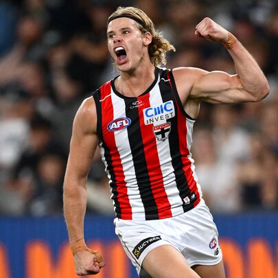 A St Kilda AFL player cheering on the field, his mouth open as if shouting, a clenched fist raised above his head, and a blurred stadium crowd behind him.