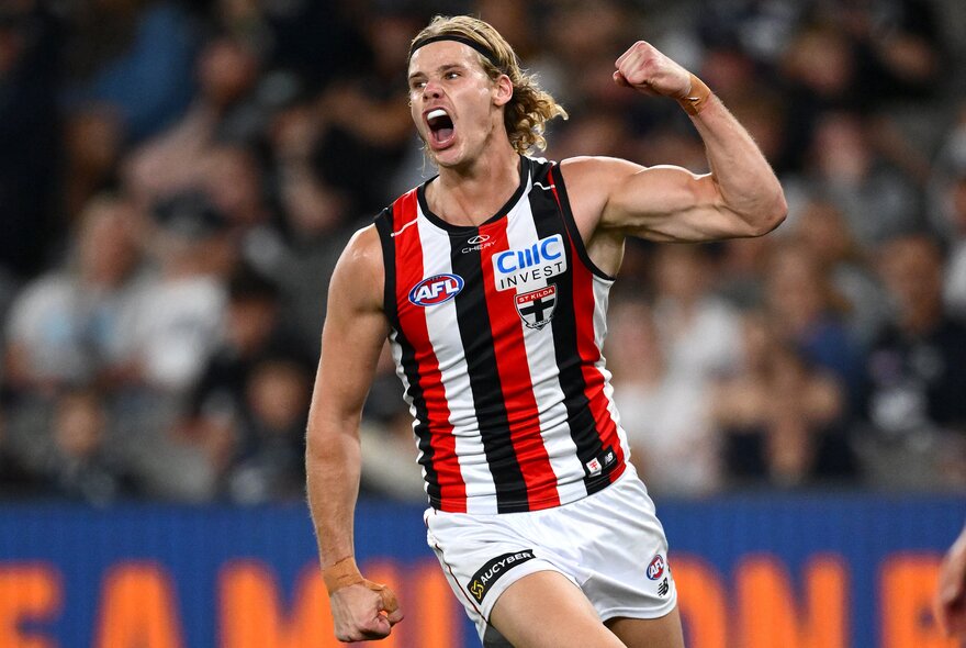 A St Kilda AFL player cheering on the field, his mouth open as if shouting, a clenched fist raised above his head, and a blurred stadium crowd behind him.