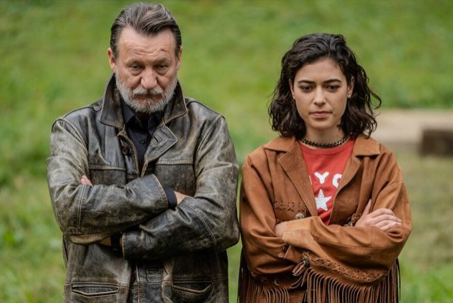 Man and woman standing with arms folded, with grass behind.