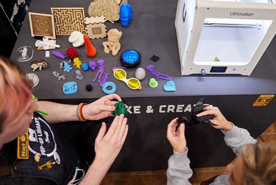 Two people holding 3D printed creations made from scrap plastic; other 3D printed creations on the table in front of them.