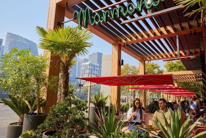 Two friends are sitting outside at a restaurant. There are large pot plants with tropical plants in front of them and a sign saying Marmont. 