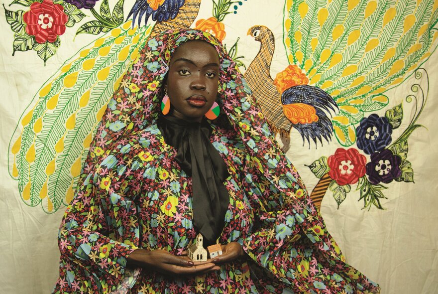 A Black woman dressed in a matching multi-coloured floral headscarf and gown, standing against a backdrop painted with green peacocks and flowers.