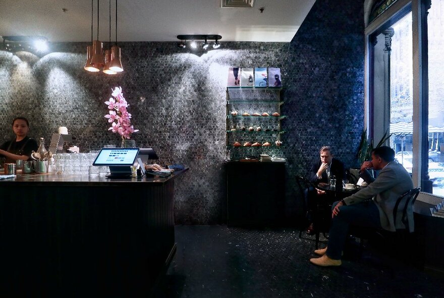 Grey tiled interior of Rosso Espresso Bar, a service counter, and two people sitting at a small window table facing the street.