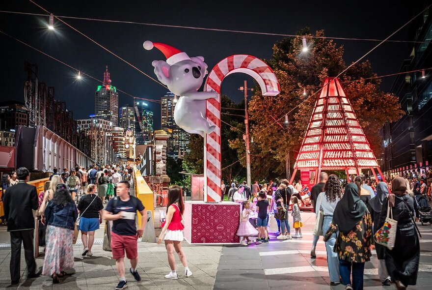 People standing around giant Christmas decorations with koalas climbing lit-up candy canes at Queensbridge Square at night, city buildings in the background.