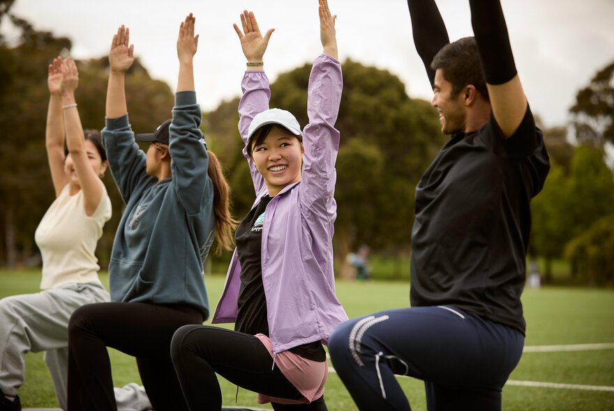 A group of four people doing an outdoor fitness or stretching class on a grassy field with trees in the background.