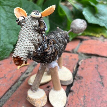 A handmade miniature creature crafted from natural materials like pinecones, twigs, and seed pods, standing on a brick surface.