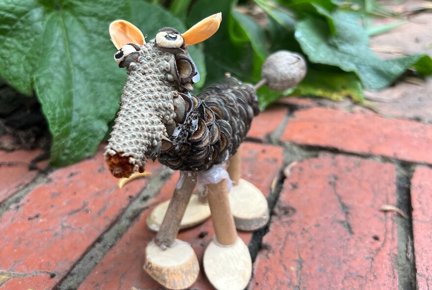 A handmade miniature creature crafted from natural materials like pinecones, twigs, and seed pods, standing on a brick surface.