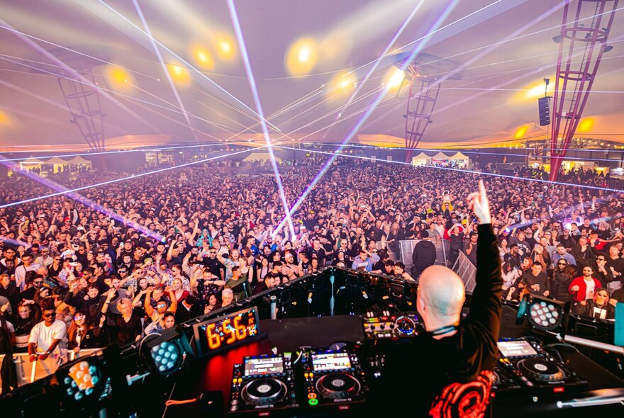 DJ at his desk with screens and controls, with huge crowd in front under lights.