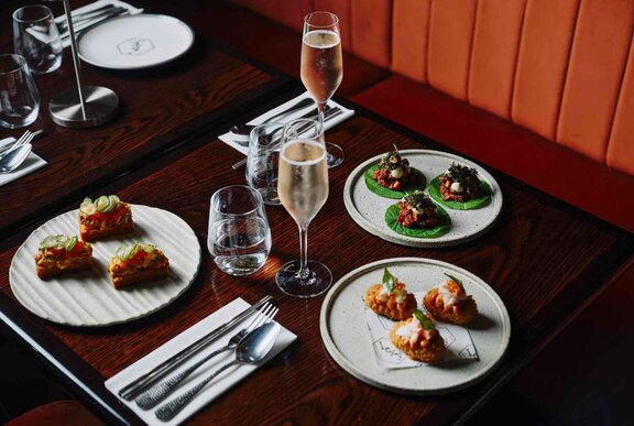 A table set for two with three small plates of canapes and two glasses of sparkling wine. 