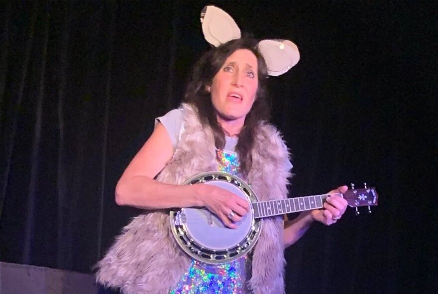 A woman dressed in a furry vest with kangaroo ears playing a banjo on stage. 