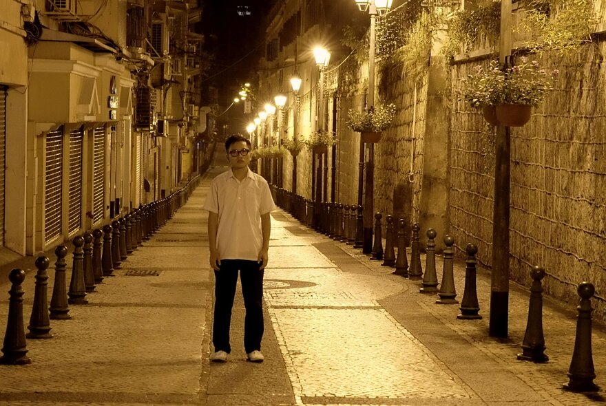 Film still of an Asian man standing in a laneway at night, lit by street lights.